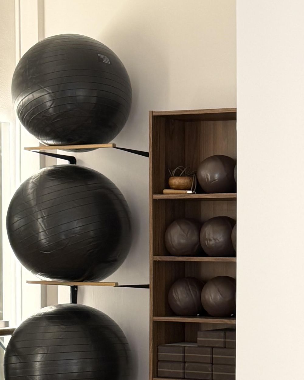 Cuatro pelotas de pilates negras apiladas en un soporte, junto a una estantería de madera con balones medicinales